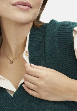 Teal gestrickter Pullover mit tiefem V-Ausschnitt. Über einem weißen Hemd mit Kragen getragen. Goldene herzförmige Halskette. Glatte Haut und natürliche Handgesten.