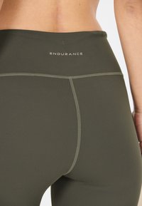 Olijfgroene sportleggings met een hoge tailleband. Voorzien van platte naden en het woord "ENDURANCE" op de achterkant gedrukt. Soepele textuur.