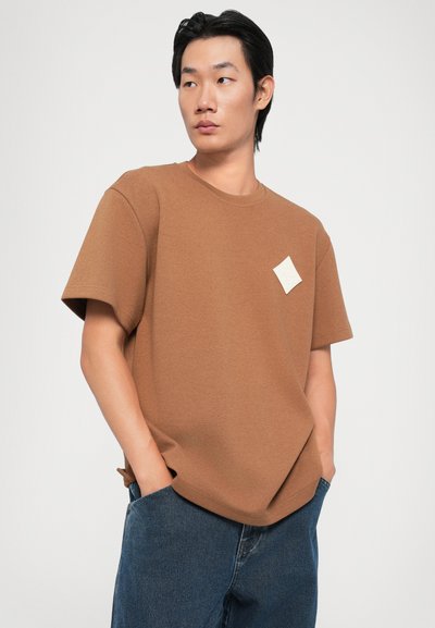 Brown short-sleeve t-shirt made of textured fabric, featuring a small white patch on the chest and a relaxed fit.