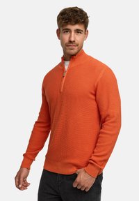 Orange tekstureret pullover med halvlukket krave, ribbede manchetter og kant. Bæret sammen med mørke jeans med tætsiddende pasform.