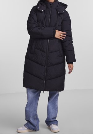 Osoba nosząca długi czarny pikowany płaszcz puchowy z kapturem, niebieskie dżinsy i białe sneakersy, stojąca na jednolitym tle.