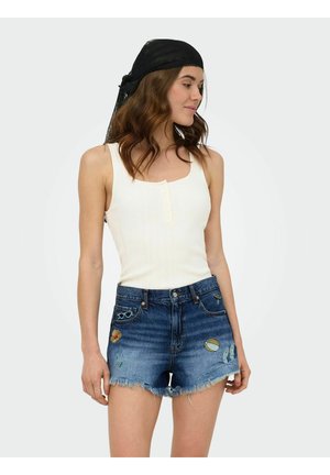 Kvinde iført en hvid ærmeløs ribbet top, denimshorts med lapper og et sort blonder hovedtørklæde, kigger til venstre.