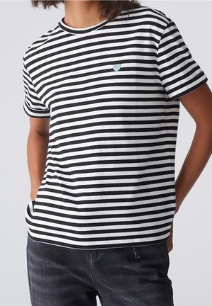 Osoba ubrana w czarno-białą, poziomo prążkowaną koszulkę z krótkim rękawem z małym emblematem serca i ciemne dżinsy, ręce w kieszeniach.