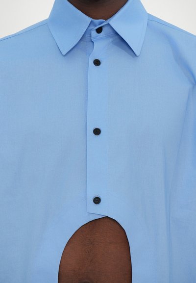 Chemise bleu clair à boutons avec un col structuré, des boutons noirs et un détail en découpe distinctif sur le bas devant. Texture lisse.
