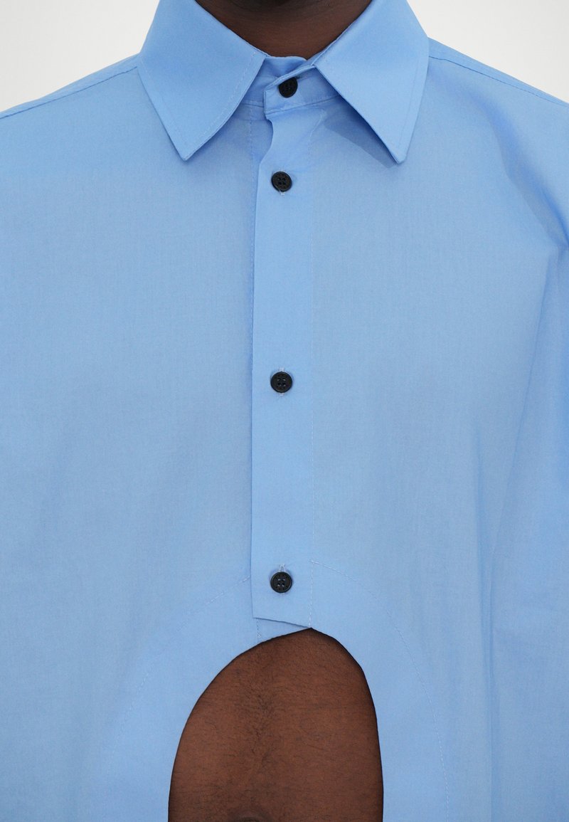 Lyseblå skjorte med opretstående krave, sorte knapper og en iøjnefaldende udskæringsdetalje på den nederste front. Glat tekstur.