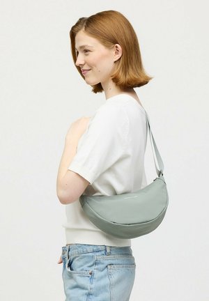 Vrouw met kort rood haar, gekleed in een wit overhemd en blauwe spijkerbroek, draagt een lichtgrijze, halvemaanvormige schoudertas.