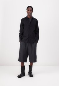 Svart knapp-up skjorte, grå løse shorts og svarte ankelstøvler med spenner. Glatt stoff, skreddersydd passform, minimalistisk design.