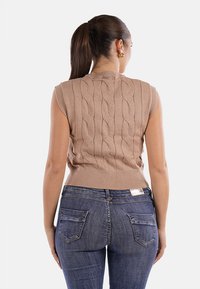 Top in maglia beige senza maniche con motivo a cavo, orlo a coste e scollo rotondo. Abbinato a jeans attillati blu scuro con tasche posteriori.