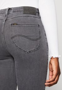 Jeans de mezclilla gris con un diseño de bolsillo trasero curvado, un parche de cuero negro texturizado y un detalle de dobladillo deshilachado en una blusa de manga larga.