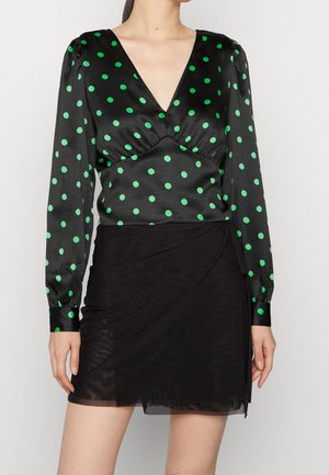 Haut en satin noir avec des pois verts, décolleté en V profond, buste froncé et manches longues. Associé à une jupe portefeuille noire transparente.