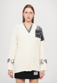 Kremno barvani pleteni kardigan z V-izrezom, debele kabelske zasnove in kontrastnimi črnimi vzorčenimi rokavi. Ima gumbe spredaj in žepke.