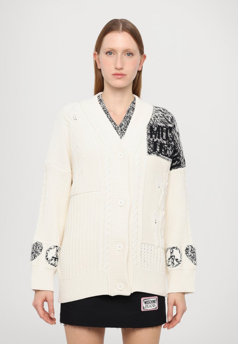 Kremno barvani pleteni kardigan z V-izrezom, debele kabelske zasnove in kontrastnimi črnimi vzorčenimi rokavi. Ima gumbe spredaj in žepke.