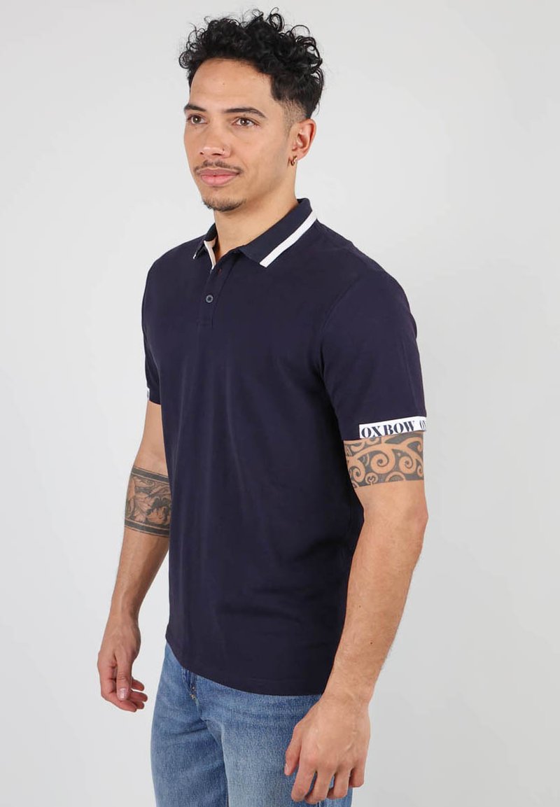 Polo marine avec col et accents de manches blancs, manches courtes et détail de logo sur la manche, associé à un jean bleu clair.