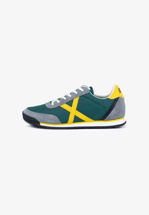 Zapatilla verde y gris con un patrón de cruces amarillas, cordones blancos, suela negra y un acento amarillo en el talón.