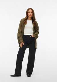 Femme debout portant une veste vert olive, un haut blanc ajusté, un jean large noir et des ballerines noires à bout pointu sur un fond blanc.