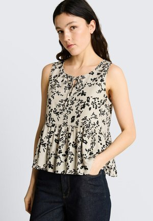 Junge Frau mit langen dunklen Haaren, die ein ärmelloses beiges Top mit schwarzem Blumenmuster und dunkle Jeans trägt, eine Hand in der Tasche.