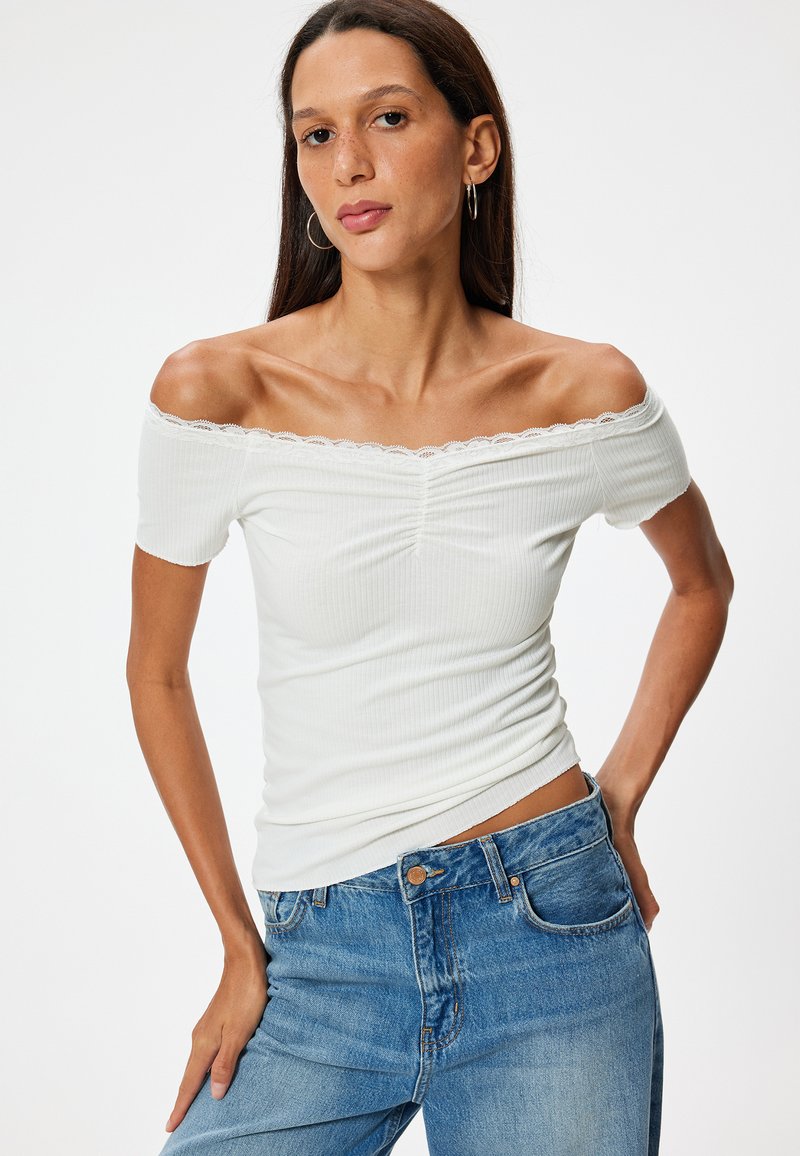 Valkoinen ribatut olkapäät paljastava paita pitsireunuksella, jossa on tiukka malli ja lyhyet hihat, yhdessä sinisten korkeavyötäröisten farkkujen kanssa.