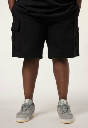 Person iført sorte cargoshorts og grå sneakers står mod en ensfarvet baggrund.