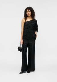 Μαύρο τοπ με έναν ώμο και διακοσμητικές φούντες, συνδυασμένο με μαύρο φαρδύ παντελόνι. Το μοντέλο κρατά μια μικρή μαύρη τσάντα και φορά μυτερά παπούτσια.