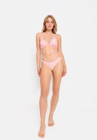 Roza bikini z geometrijskim vzorcem, zgornji del v obliki trikotnika s križnimi naramnicami in ujemajoče se hlačne s stranskimi izrezi. Gladka tekstura.