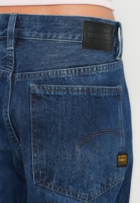 Σκούρο μπλε τζιν με ελαφρώς φθαρμένη υφή, που διαθέτει μαύρο δερμάτινο πίσω σημείο και κίτρινη ετικέτα μάρκας στην δεξιά τσέπη.