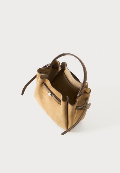 Petit sac à main en daim beige avec poignée en cuir marron foncé et brides latérales, ouverture sur le dessus et intérieur visible sur un fond clair uni.