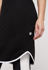 Svart klänning med en böljande nederkant, vita detaljer och en liten logotags. Slät tygtextur med en åtsittande silhuett.