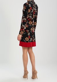 Žena oblečená v černých květinových šatech s červeným lemem a nahých lodičkách, stojící zády k fotoaparátu.