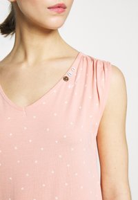 Top senza maniche di un tenue rosa, con scollatura a V, decorato con piccoli fiori bianchi, tre bottoni vicino alla spalla e sottili dettagli arricciati.