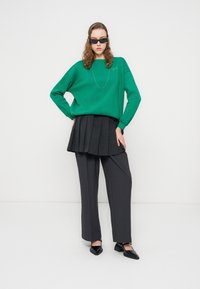 Πράσινο φούτερ με μακριά μανίκια, συνδυασμένο με σκουρόχρωμη πλισέ φούστα και παντελόνια με ίσια γραμμή. Μαύρα παπούτσια με μυτερό σχήμα.