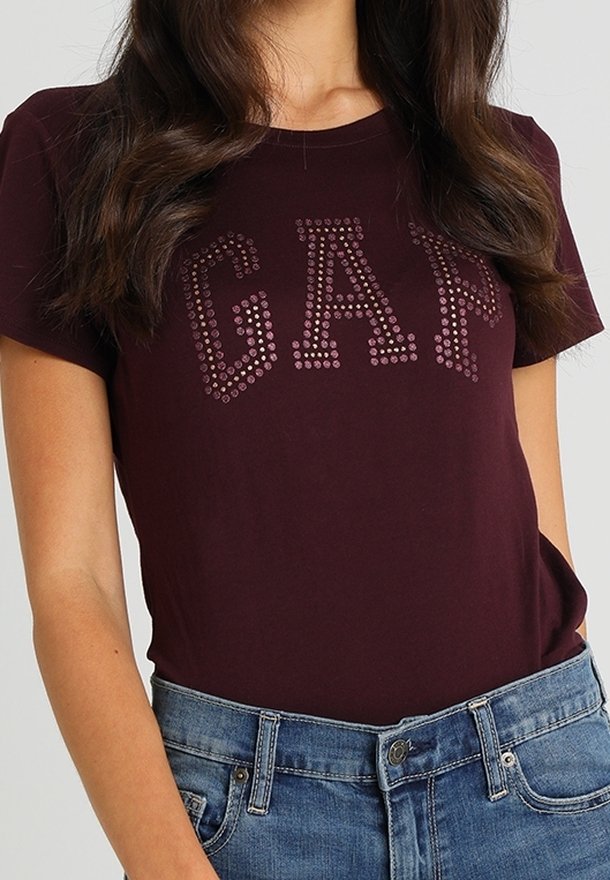 Femme portant un t-shirt à manches courtes bordeaux foncé avec "GAP" en petits clous ronds, associé à un jean en denim bleu.