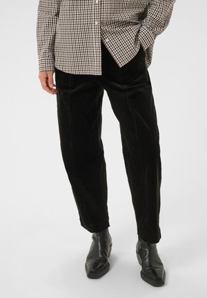Personne portant un pantalon en velours côtelé noir, des bottines en cuir noir et une chemise à carreaux à manches longues, une main dans la poche.
