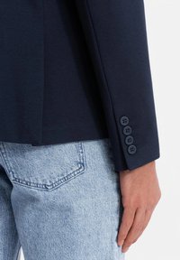 Marineblazer mit glatter Textur, ausgestattet mit vier Knopfverschlüssen am Ärmel. Kombiniert mit hellblauen Jeans.
