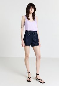 Donna con capelli mossi e bagnati, che indossa un top senza maniche lavanda, pantaloni corti in denim scuro a vita alta e sandali neri con tacco, in piedi su uno sfondo bianco.