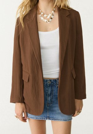 Vrouw draagt een bruine blazer over een wit hemdje, gecombineerd met een blauwe denim minirok en gelaagde gouden muntenkettingen.