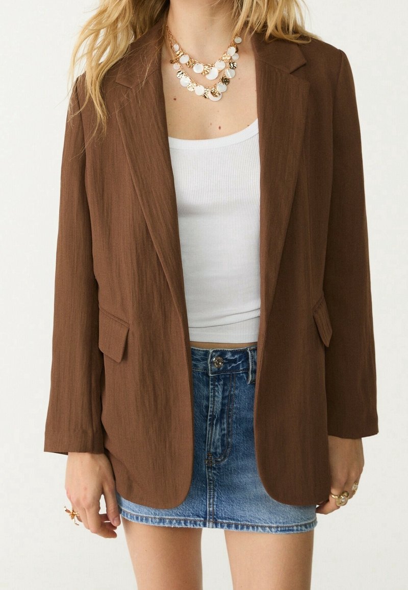 Femme portant un blazer marron sur un débardeur blanc, associé à une mini-jupe en jean bleue et des colliers superposés en pièces d'or.