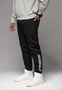 Pantalon noir fuselé avec des bandes blanches sur les côtés, porté avec un sweat-shirt gris et des baskets blanches montantes. Tissu lisse et coupe ajustée.