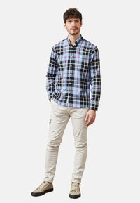 Camisa de cuadros en negro, azul y rojo, con cuello extendido y mangas largas. Combinada con pantalones de color beige claro y zapatos.