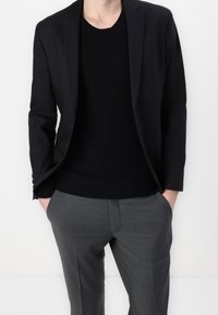 Svart kavaj över en svartsittande tröja, matchad med grå skräddarsydda byxor. Kavajen har en slitskrage och subtila axelvaddar.