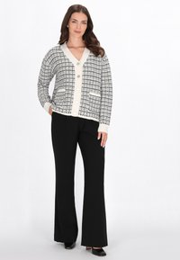 Fekete-fehér mintás kardigán V-nyakkivágással, gombos záródással és zsebekkel. Fekete nadrággal és magassarkúval párosítva. Sima, kötött textúra.