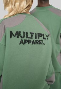 Zwei Personen tragen grüne und graue Sweatshirts, wobei eines auf dem Rücken mit dem schwarz gestickten Text "MULTIPLY APPAREL" versehen ist.