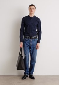 Navy button-up shirt, blue jeans, black belt, and shoes. Holding a black tote bag. Slim fit design with a smooth fabric texture.
