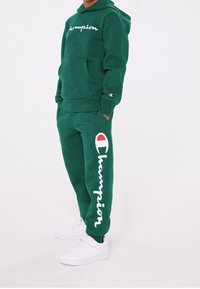 Grön fleecehuva och joggers-set med vit logotext och detaljer. Ett par vita sneakers fulländar outfiten.