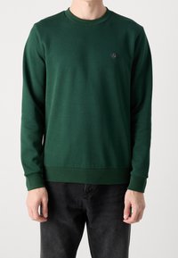 Felpa verde scuro realizzata in tessuto liscio, con scollo rotondo, maniche lunghe e un piccolo logo sul petto, abbinata a jeans neri.