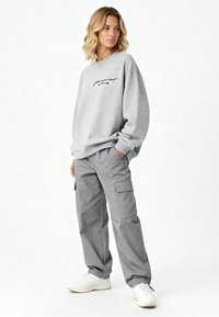 Sweat-shirt gris avec texte brodé noir et épaules tombantes, associé à un pantalon cargo gris avec poches latérales et ourlets élastiques. Baskets blanches.