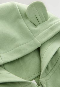 Hoodie vert clair avec une capuche arrondie et un détail en relief représentant une oreille. Le tissu est doux, confortable et présente un motif de couture visible.