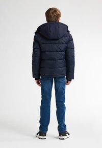 Jachetă puffer marină cu glugă, având un design matlasat și manșete ribbate, asortată cu blugi albaștri și adidași negri.