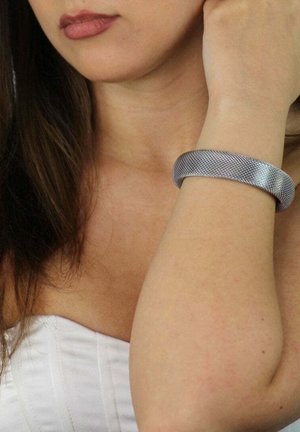 Bracciale in rete argentata con bordi lisci e arrotondati, caratterizzato da un motivo testurizzato. Indossato su un polso contro la pelle nuda.