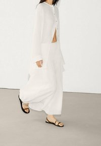 Hvid linnedskjorte med knapper, lagdelt over en hvid plisseret maxinederdel. Sorte flade sandaler. Model går mod en neutral baggrund.