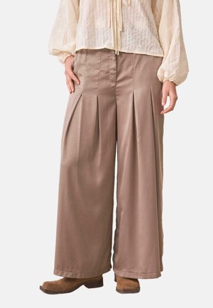 Pantalones anchos color topo con pliegues frontales, combinados con una blusa crema bordada con flores y botas de ante marrón sobre un fondo blanco.